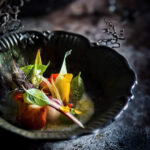 Obra fotográfica que mezcla lo culinario con lo simbólico. Un plato de pollo sobre un fondo de plumas que evoca el viaje, la libertad, y también la ironía. Juego visual que transforma lo cotidiano en arte.